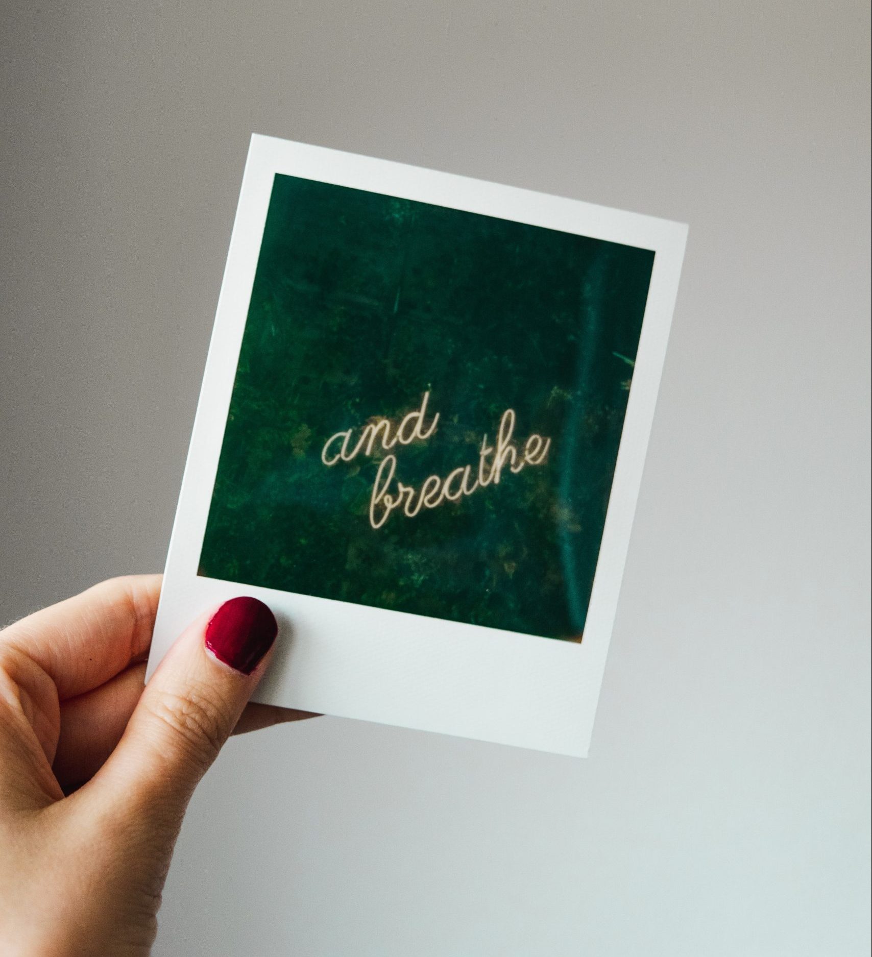 mano di donna con smalto rosso che regge polaroid con scritta a neon su fondo verde "and breath"
