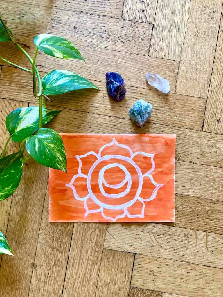 simbolo del secondo Chakra colorato di arancione e bianco, con cristalli e una pianta appoggiati su pavimento in parquet